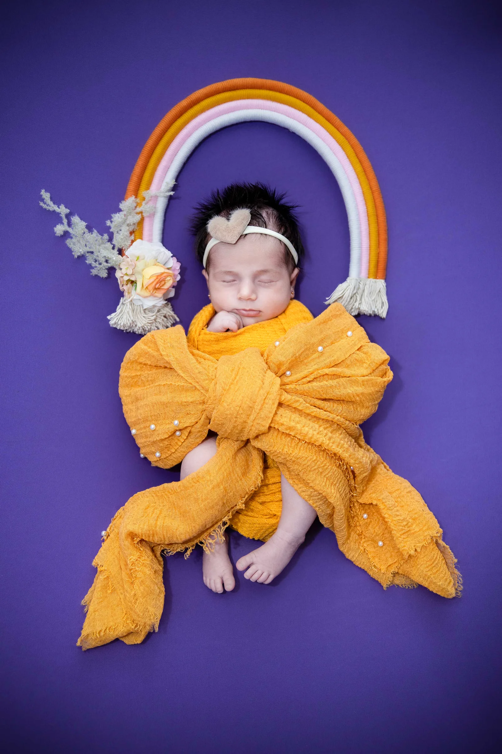 Newborn wrapped in a cozy mustard wrap beneath a gentle pastel rainbow.