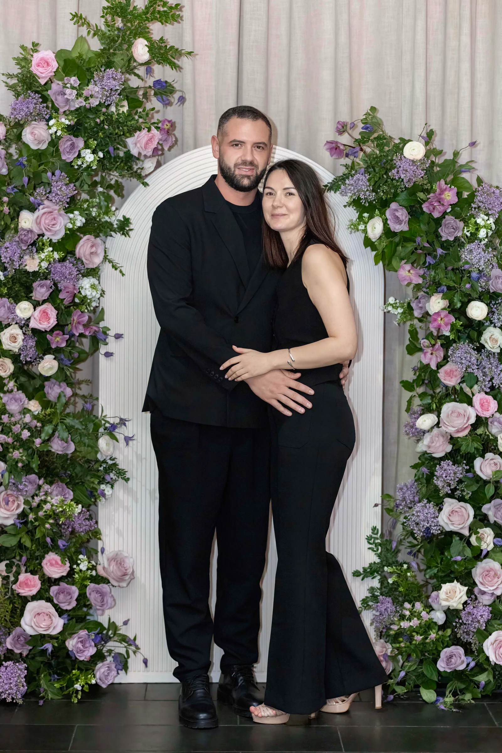 Couple in black attire sharing a tender embrace against a romantic floral backdrop.