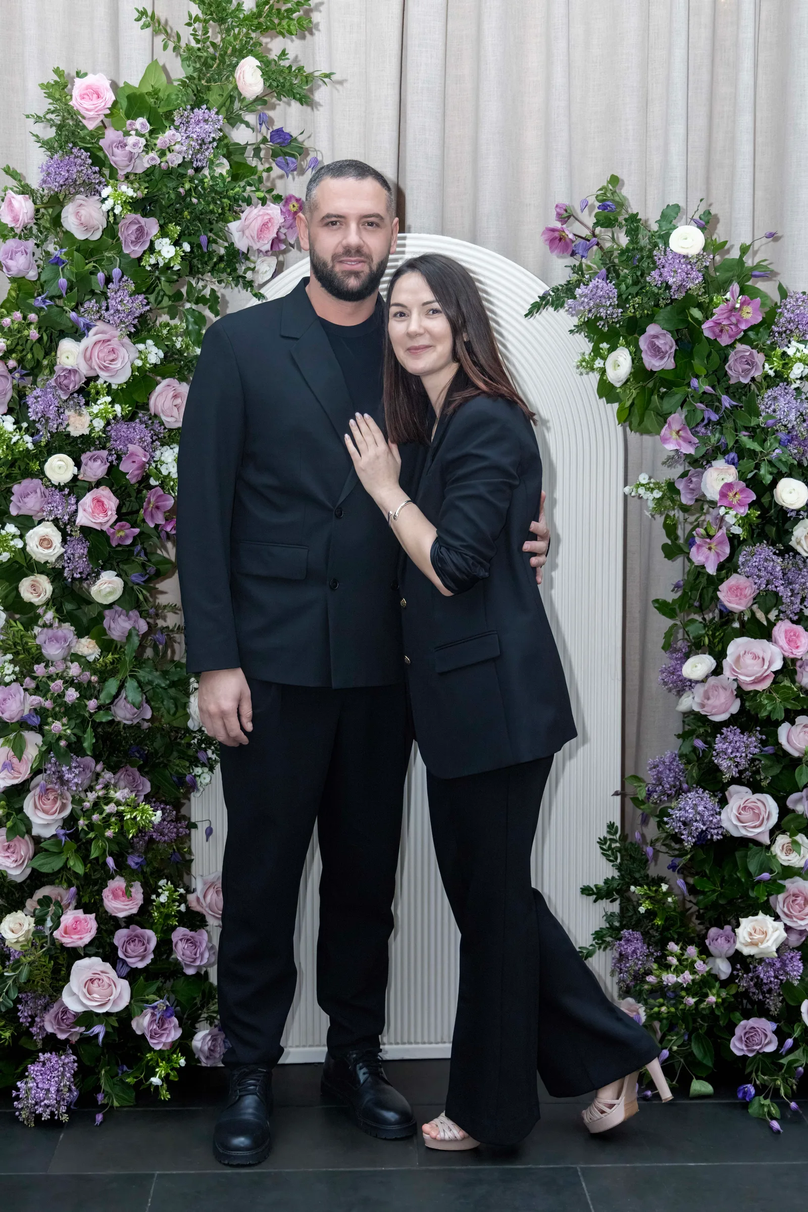 Elegant couple in black, posing affectionately beside a floral arch.