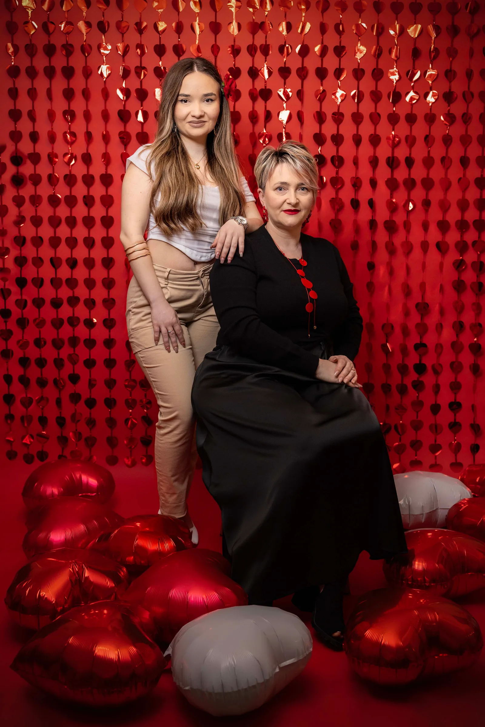 Two generations posing together against a shimmering red backdrop, surrounded by heart-shaped balloons.