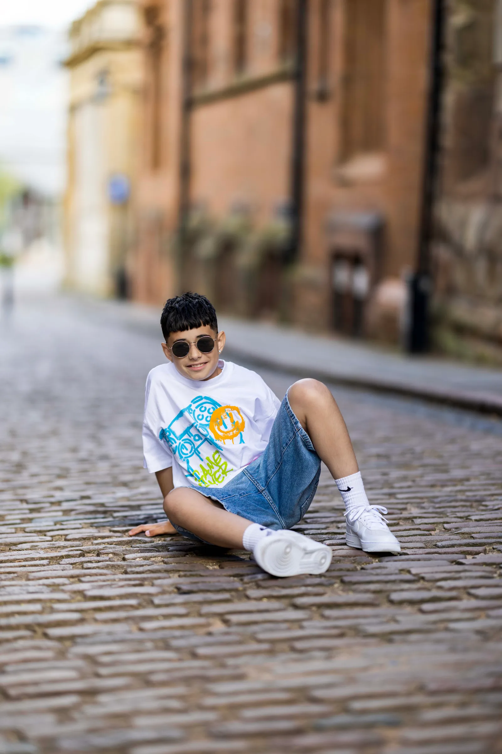Young boy in sunglasses sitting casually on a cobblestone street