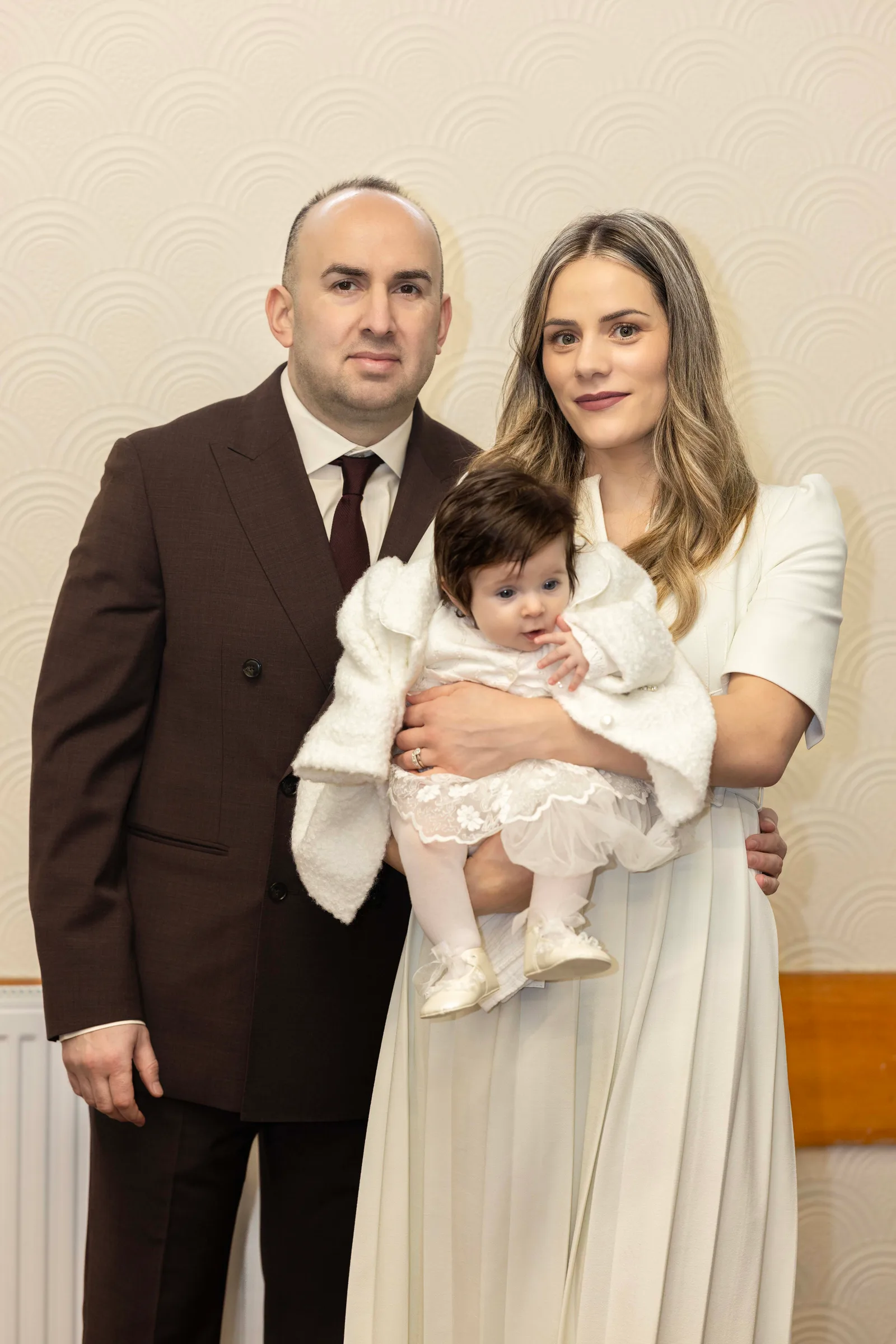 Parents holding their baby girl in her christening gown, captured in a warm, emotional moment.