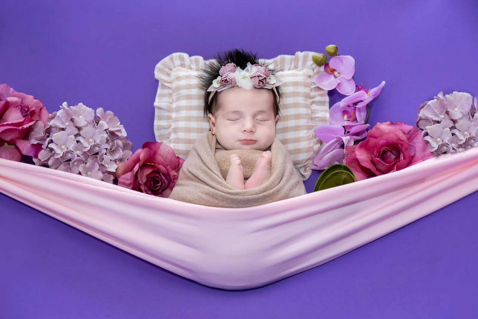 Peaceful newborn cradled in a soft pink hammock, surrounded by delicate roses and orchids.