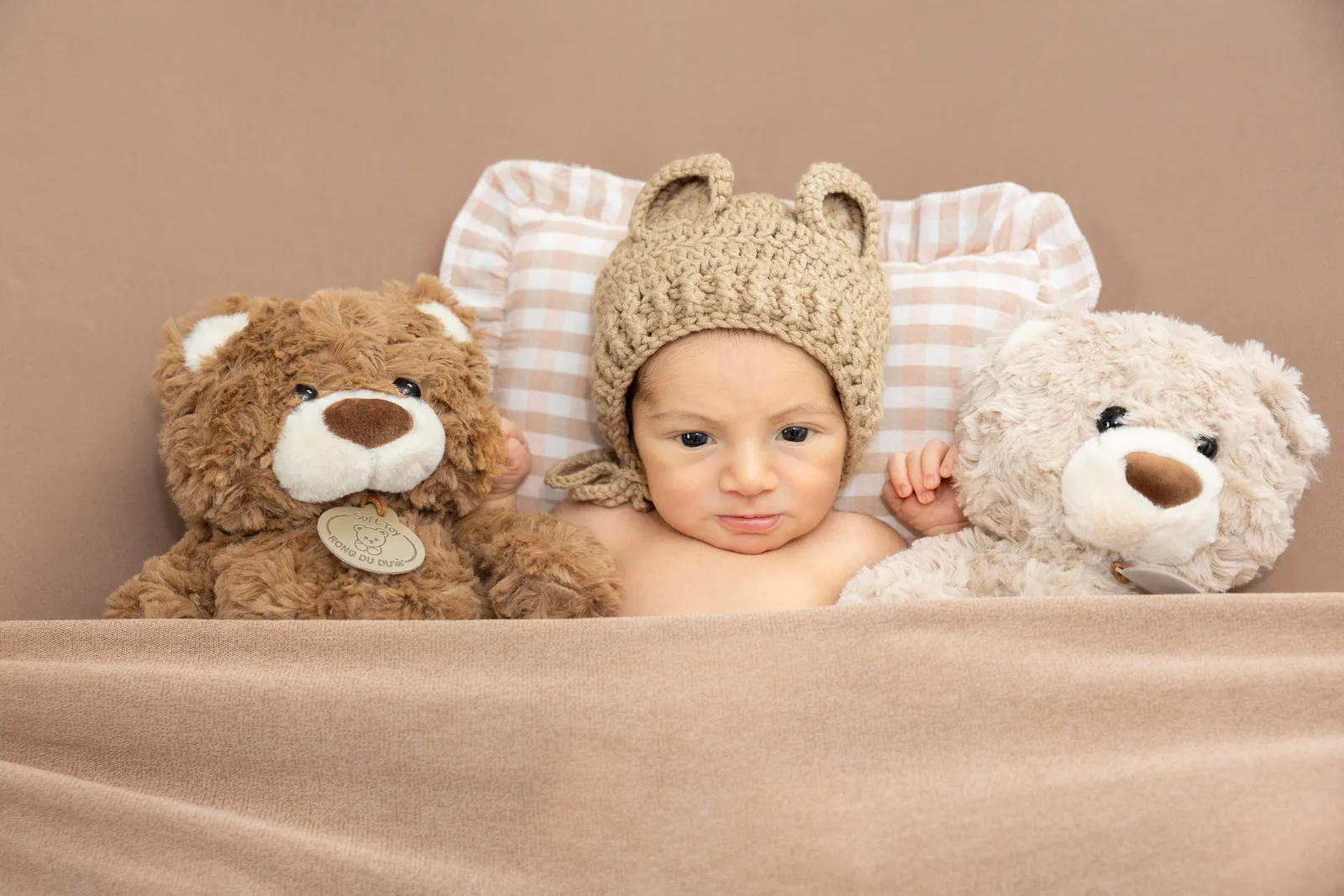 Baby wearing a brown bear-ear bonnet among a teddy bear and pink gingham pillows