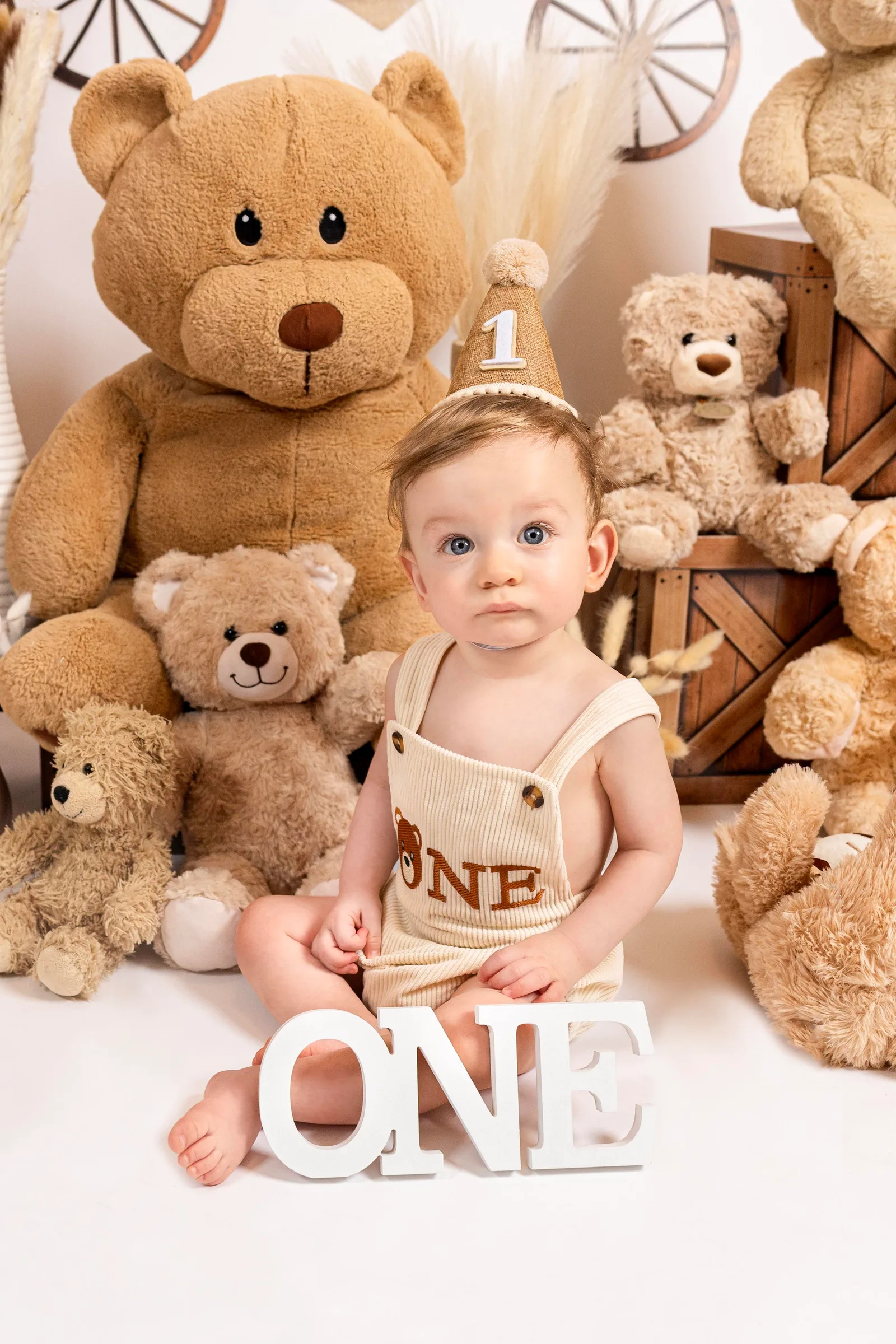 Baby in a birthday hat sitting beside a large teddy bear with wooden ONE letters