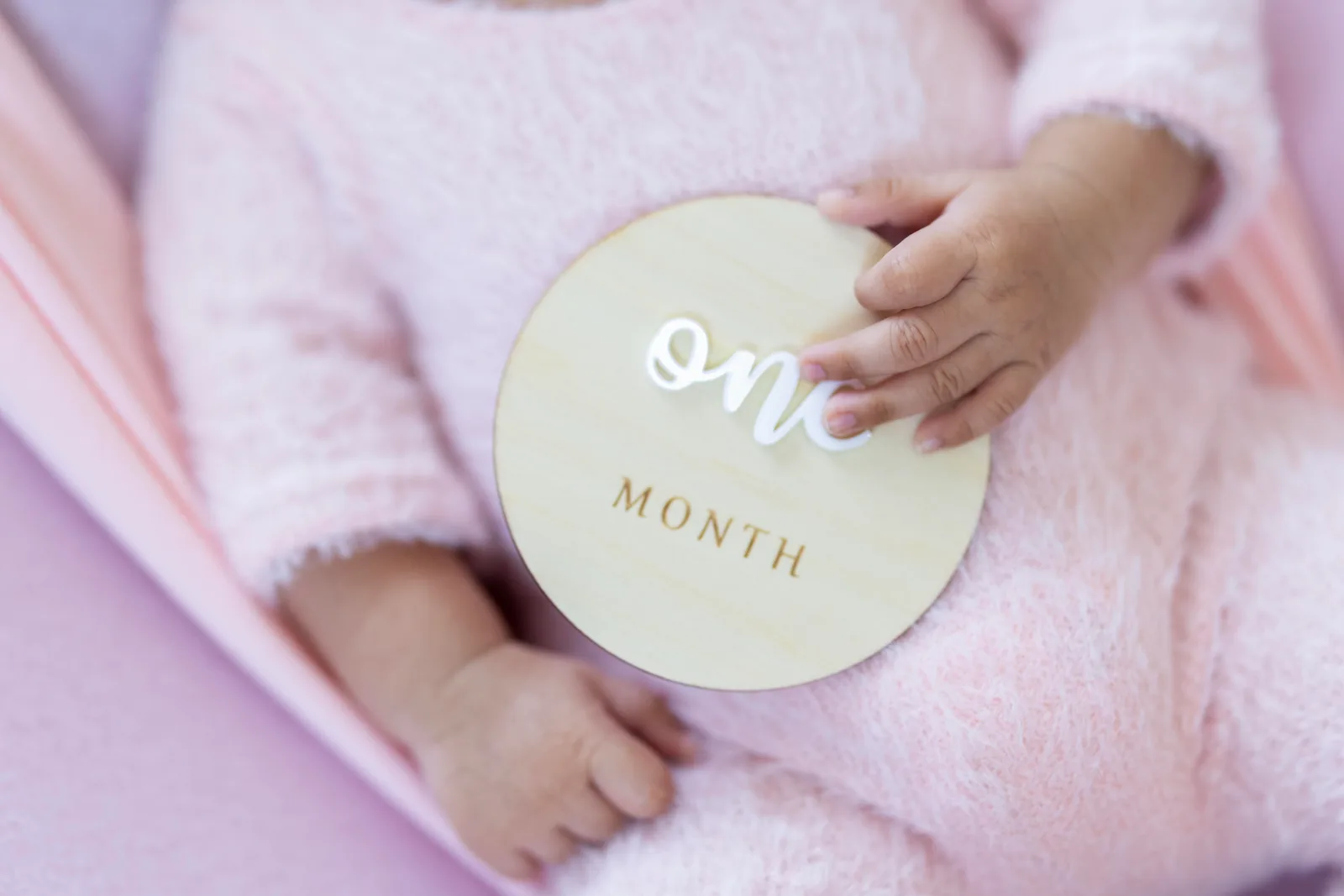 Adult hand holding a 'one month' milestone card beside a baby wrapped in pink