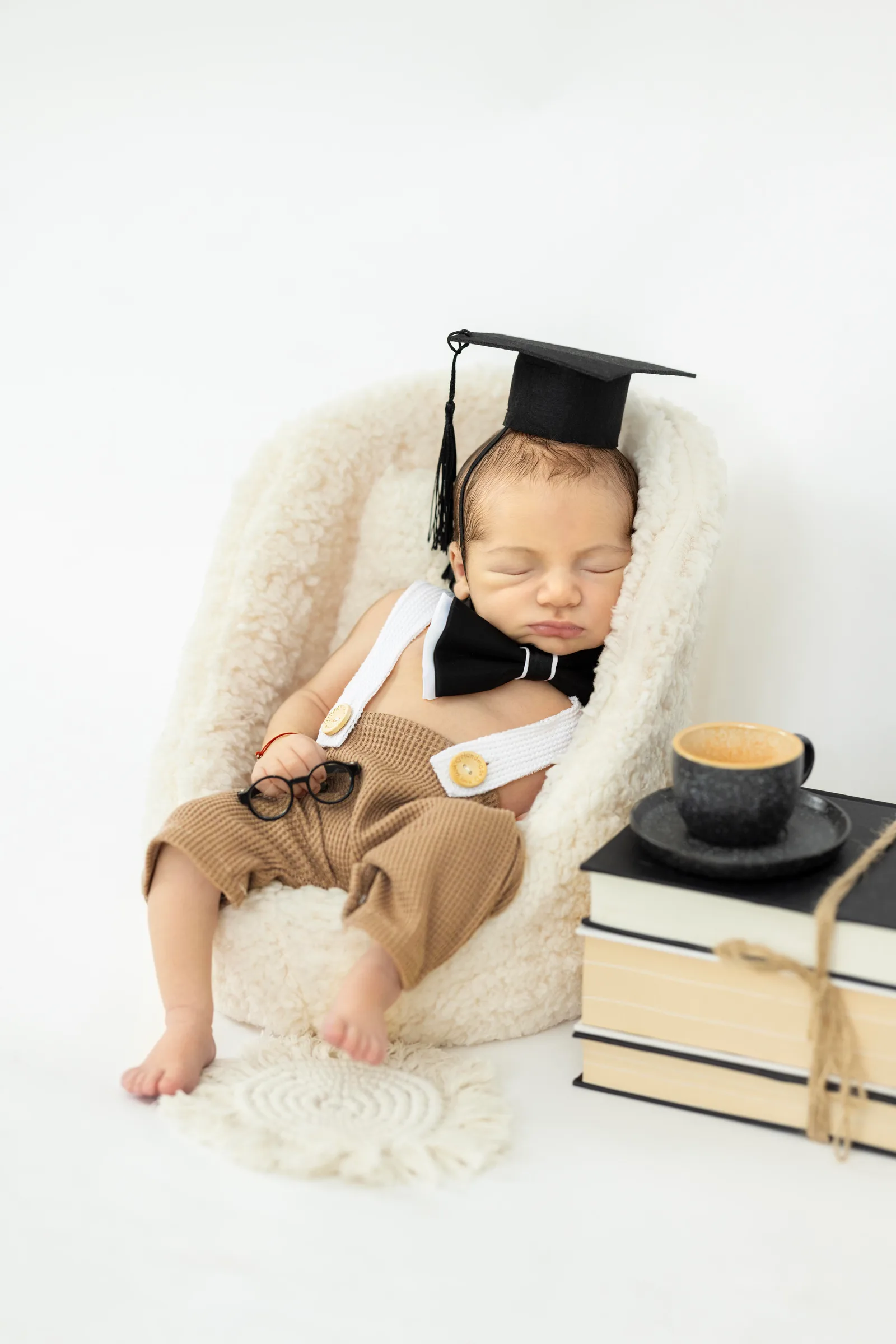 Newborn asleep in a fuzzy basket wearing a graduation cap, glasses, and bow tie beside a stack of books