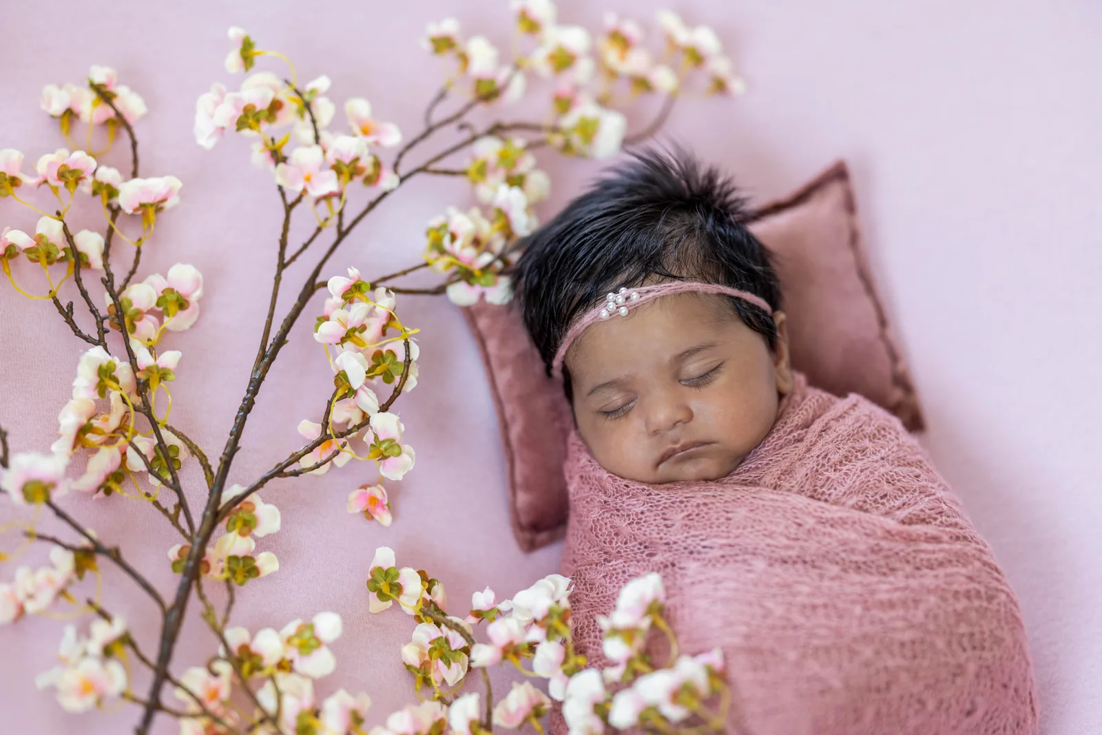 Newborn in pink wrap and pearl headband sleeping on a pink cushion among blossoms