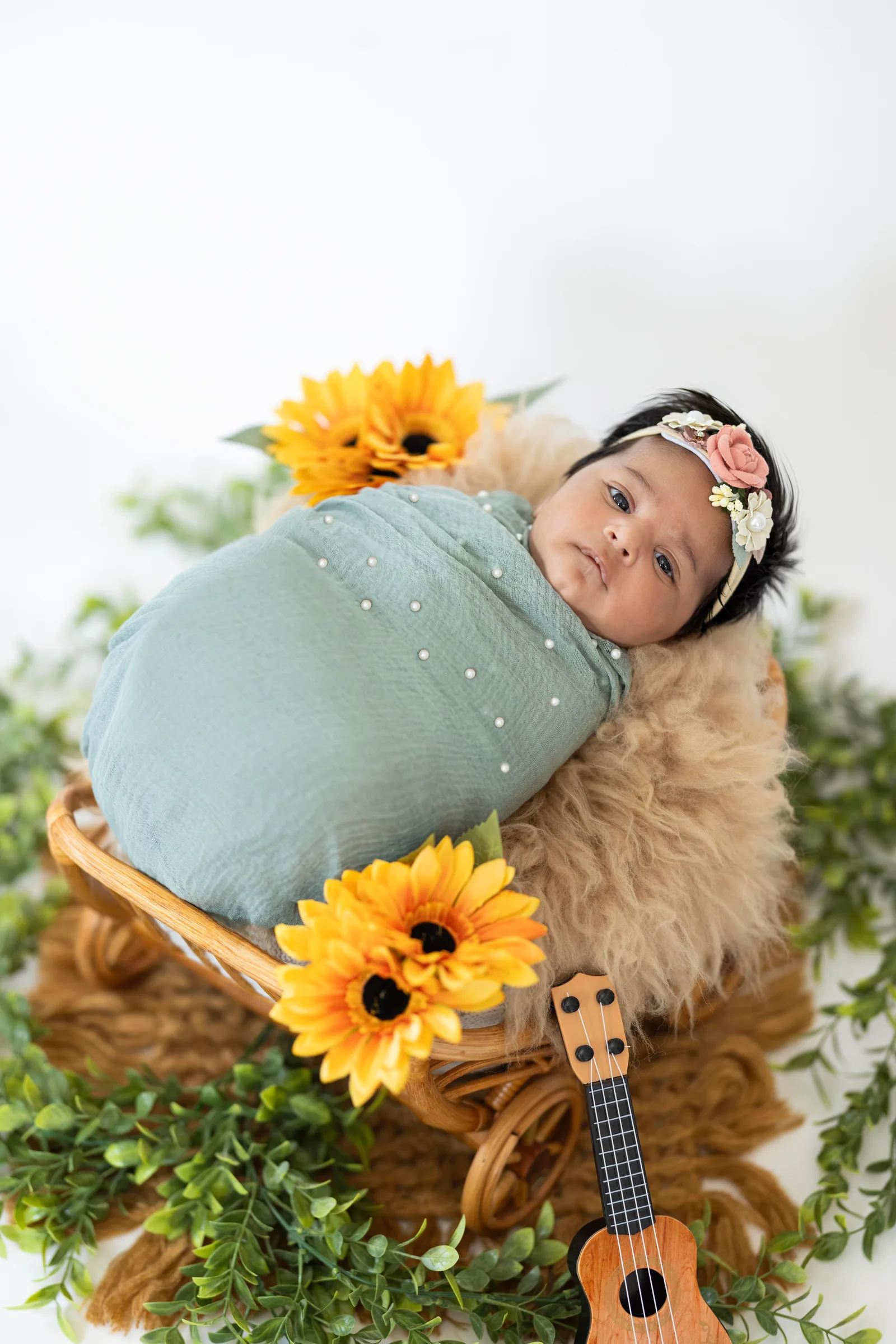 Baby in a teal wrap and floral headband resting in a basket surrounded by sunflowers