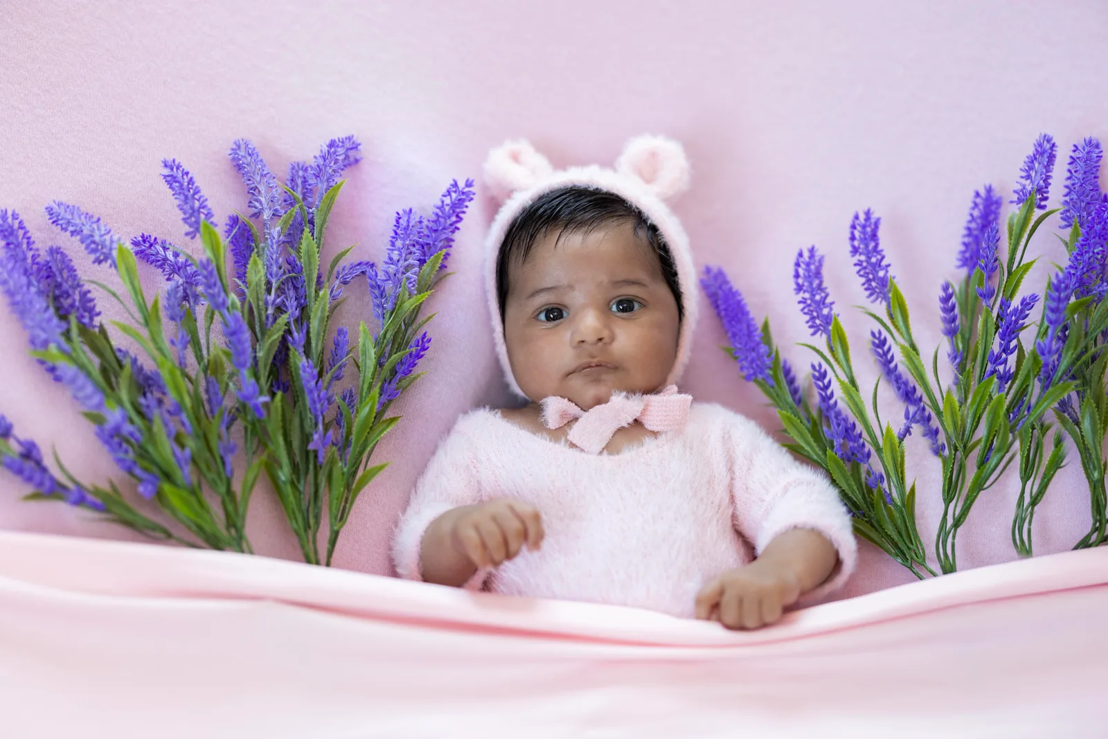 Baby wearing a white bear-ear bonnet with pink bow among lavender sprigs