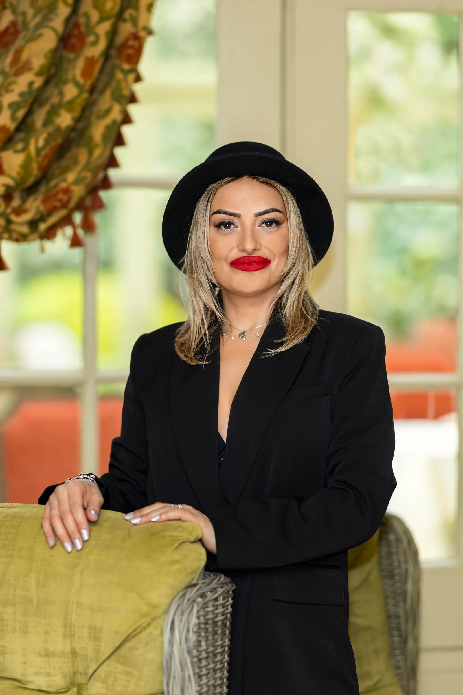 Woman in a black blazer and wide-brim hat posing near a bright window with greenery