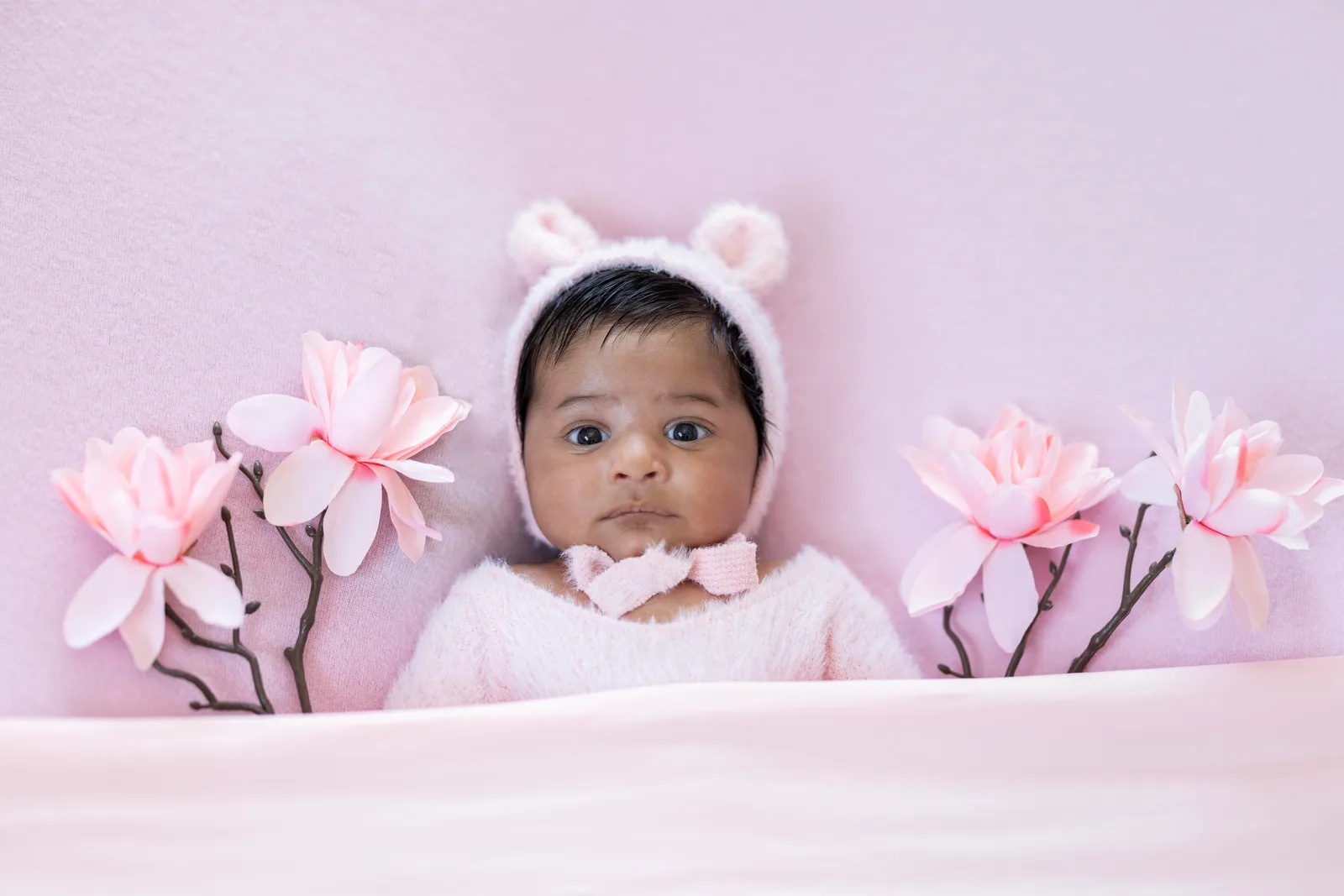 Baby wearing a white bear-ear bonnet with a pink bow among paper flowers