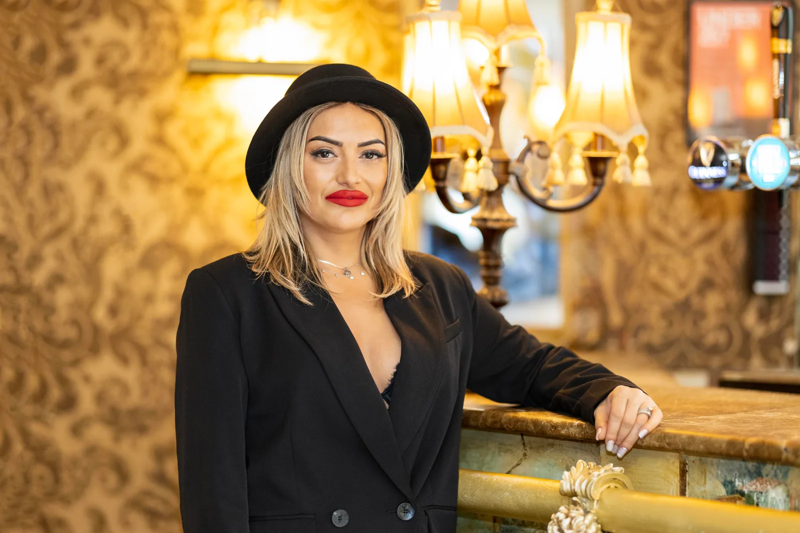 Woman in a black blazer and wide-brim hat posing in an ornate gold-toned interior