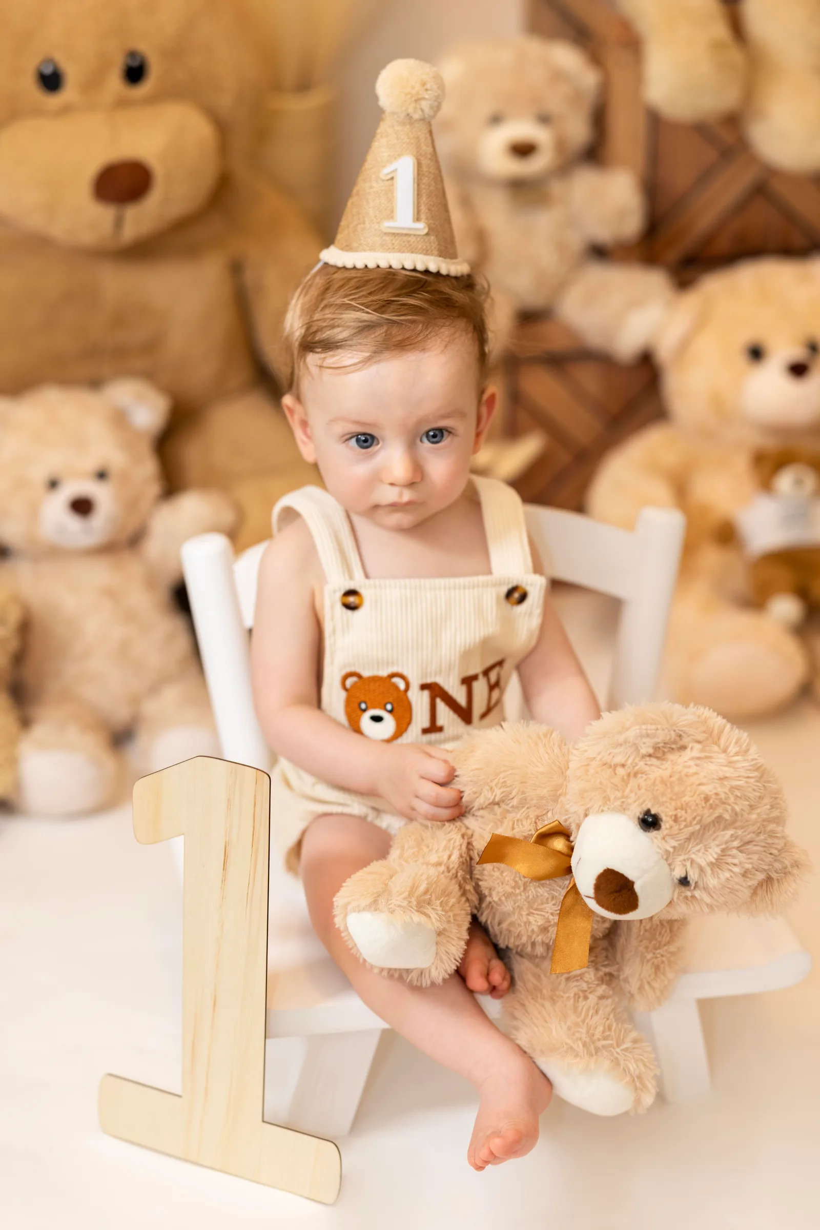 Baby wearing a birthday hat and 'ONE' overalls, sitting in a chair holding a teddy bear