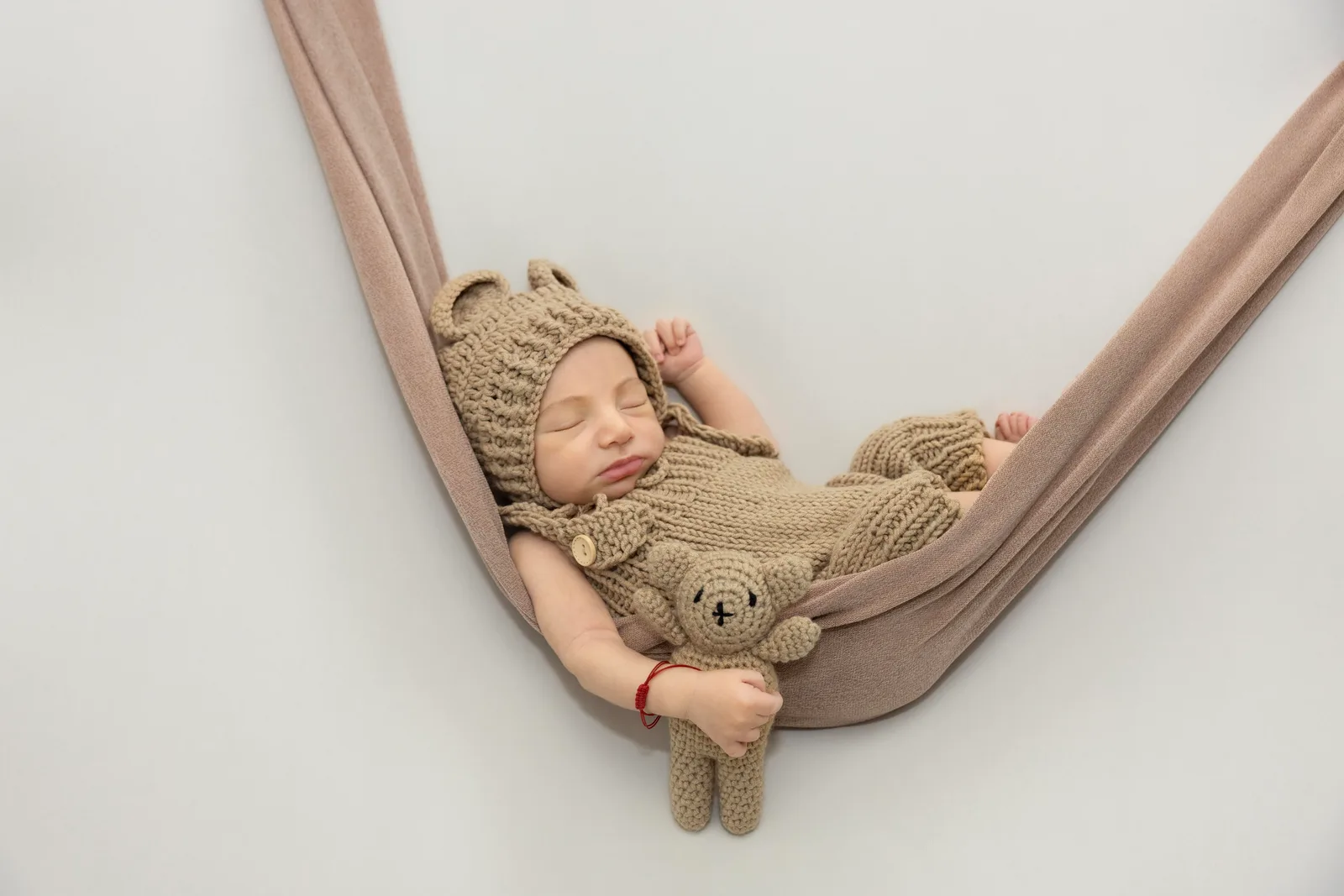 Newborn asleep in a knitted brown hammock wearing a bear hat and holding a teddy bear