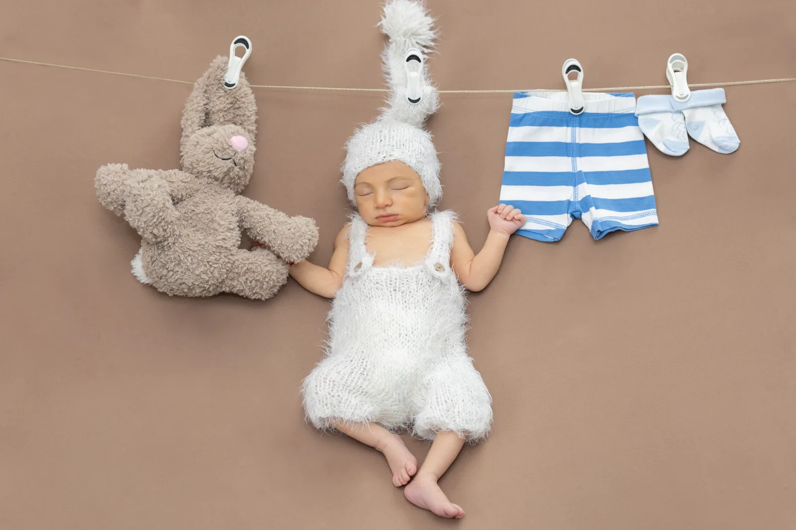 Newborn dressed as a bunny appearing to hang from a clothesline beside a stuffed bunny