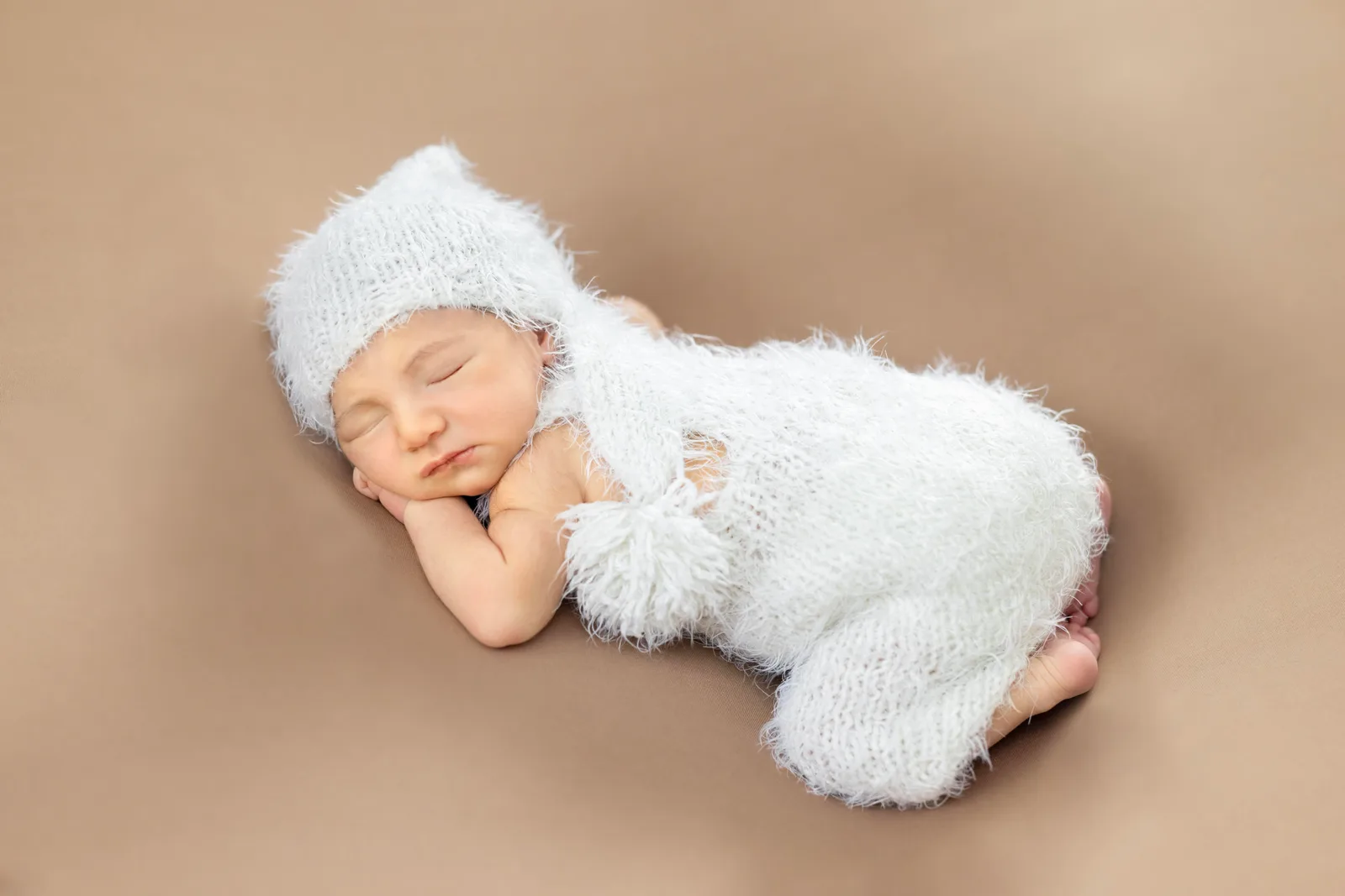 Newborn asleep in a fuzzy white knitted hat and romper on a beige backdrop