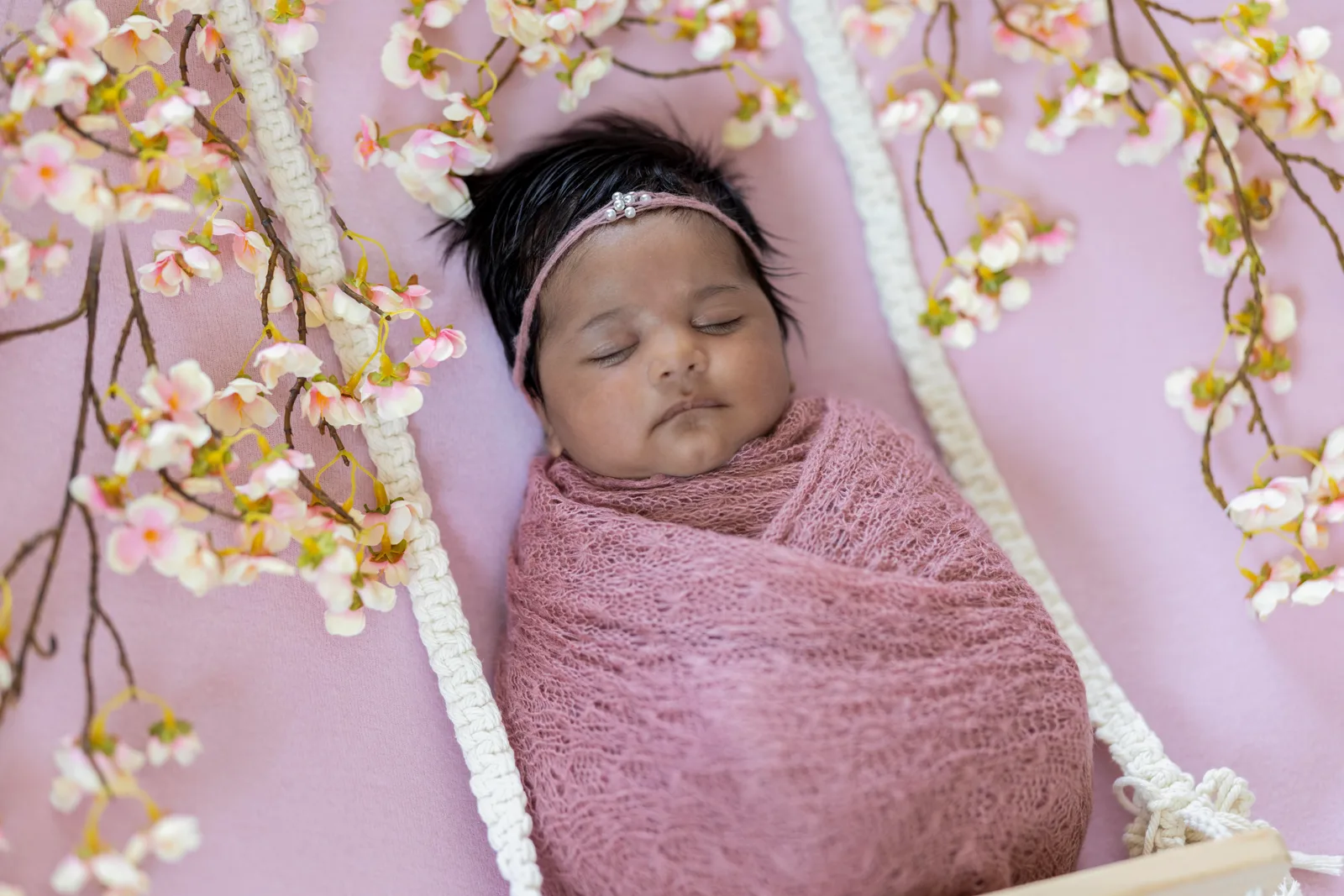 Newborn wrapped in pink knit with pearl headband among cherry blossom branches