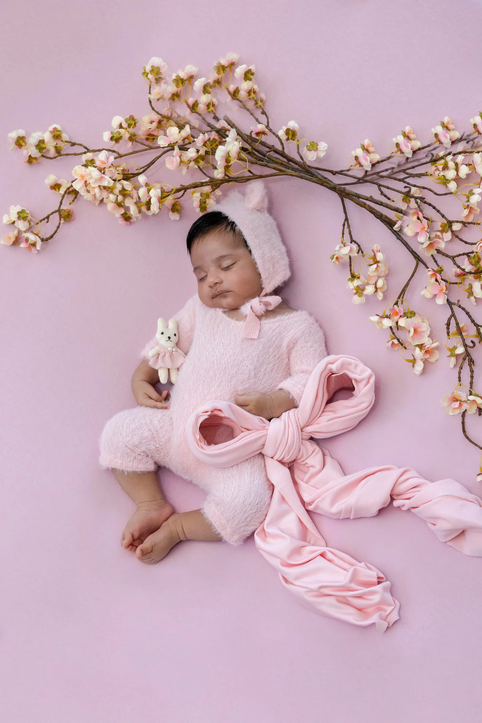 Newborn baby asleep in a pink bear bonnet and outfit, surrounded by cherry blossoms