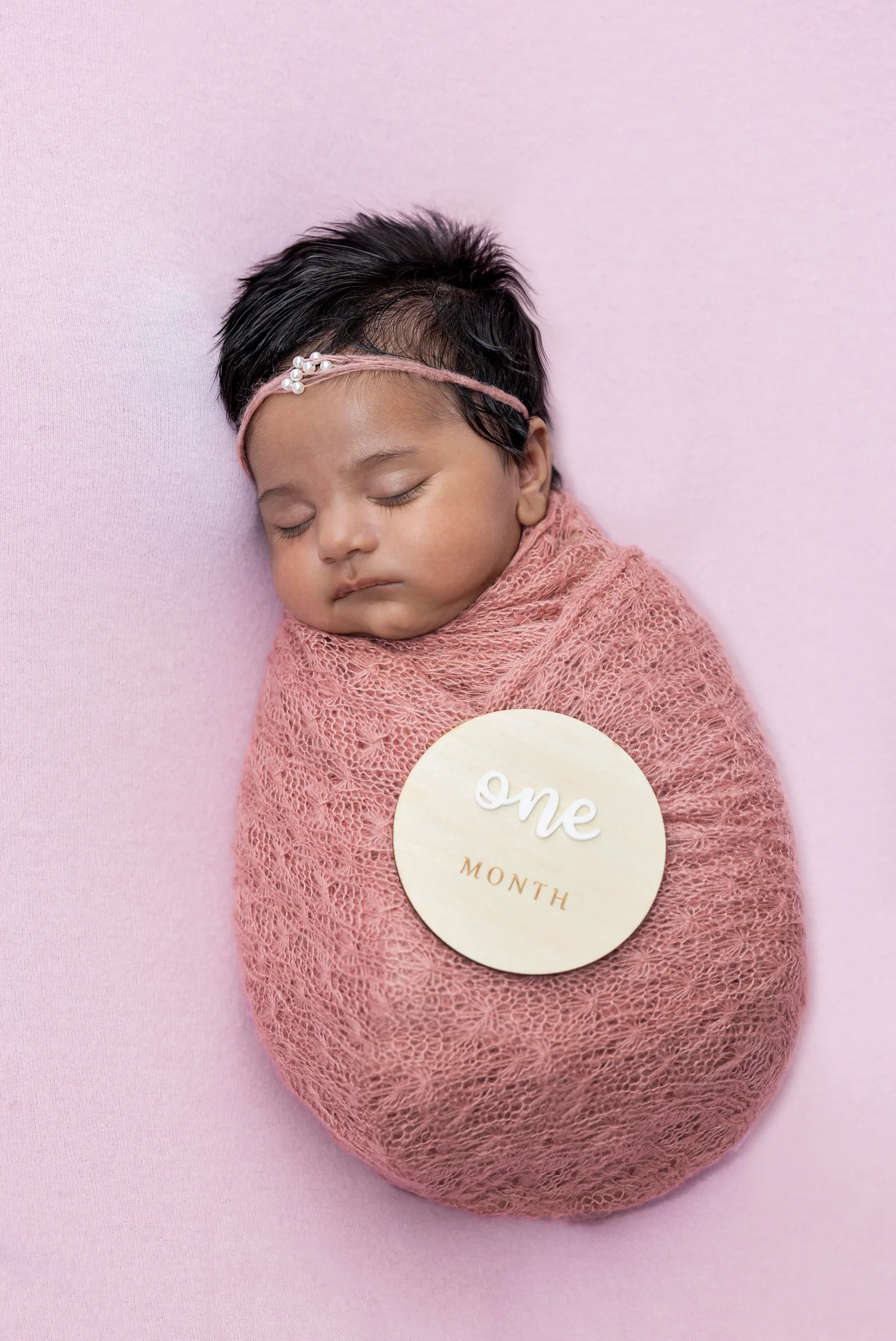 Newborn baby swaddled in dusty pink wrap with a beaded headband and one-month milestone marker
