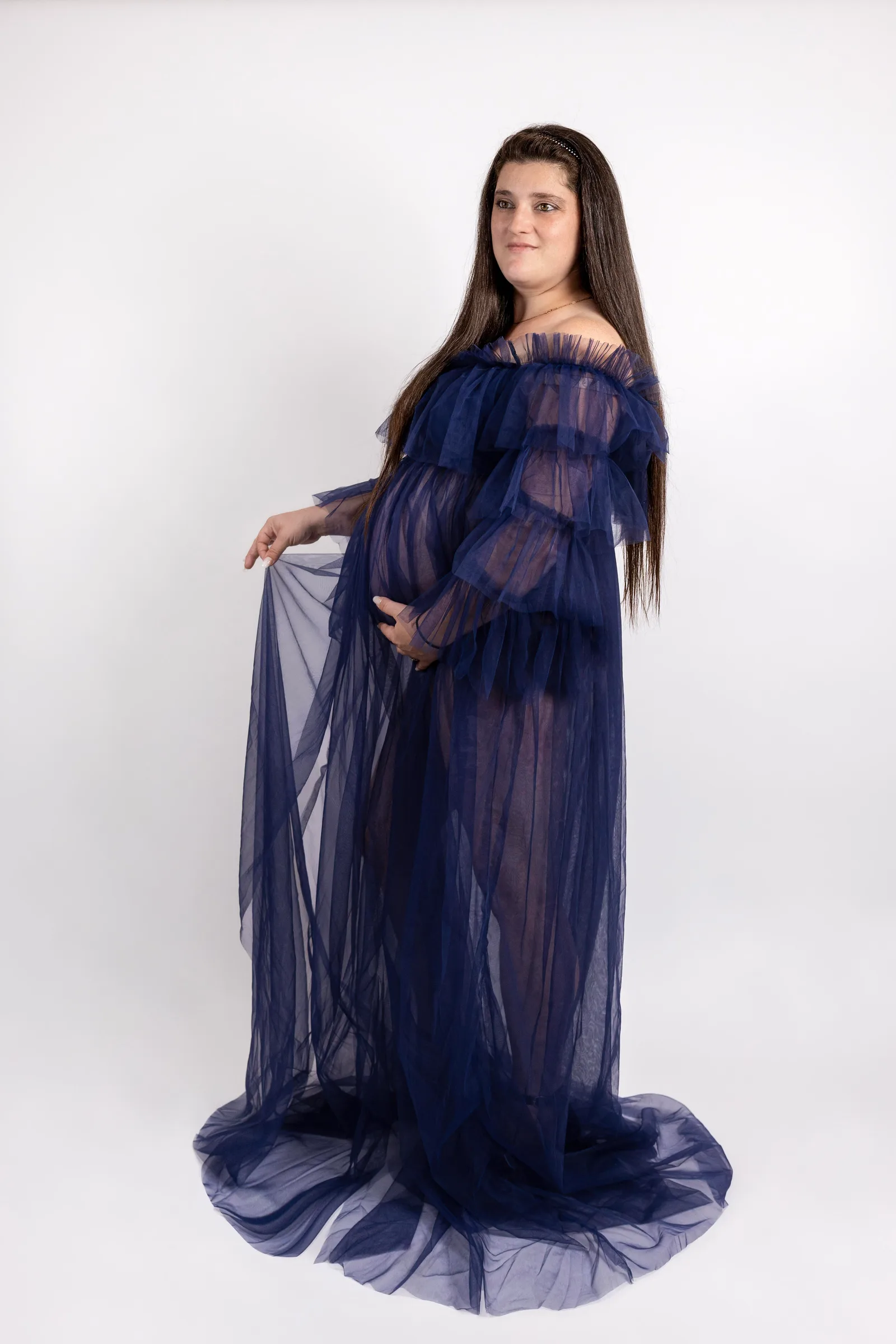 Pregnant woman standing in sheer navy tulle gown, holding the flowing train to one side