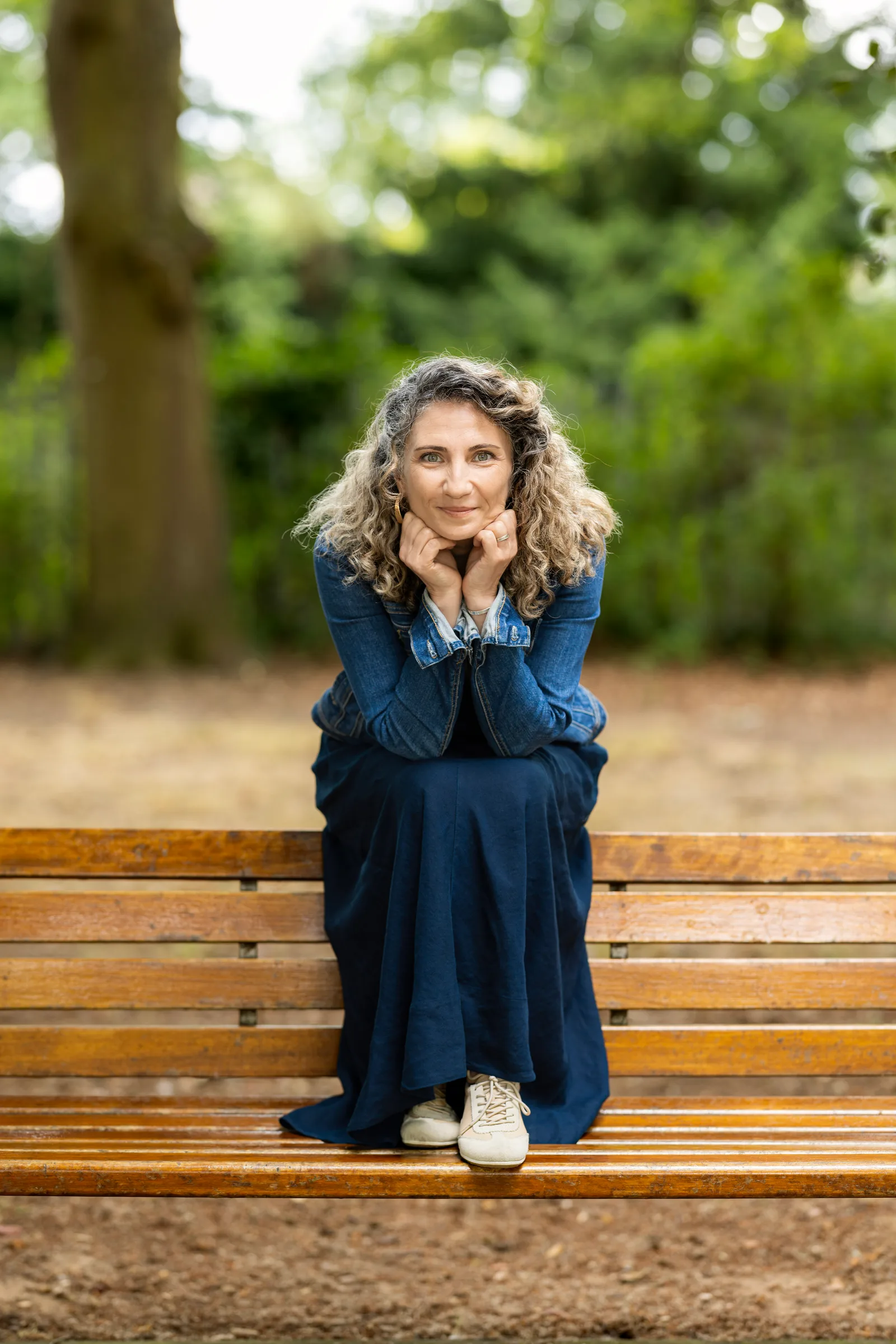 Woman in navy dress and denim jacket sitting on a park bench, hands under chin