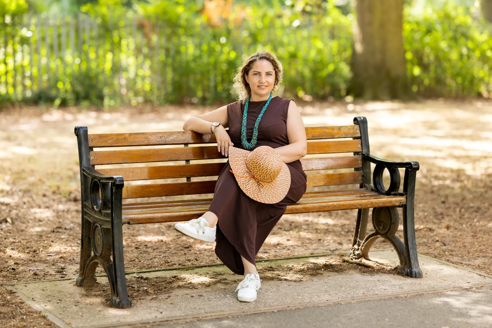 Woman sitting on a park bench in a brown dress, holding a straw sun hat
