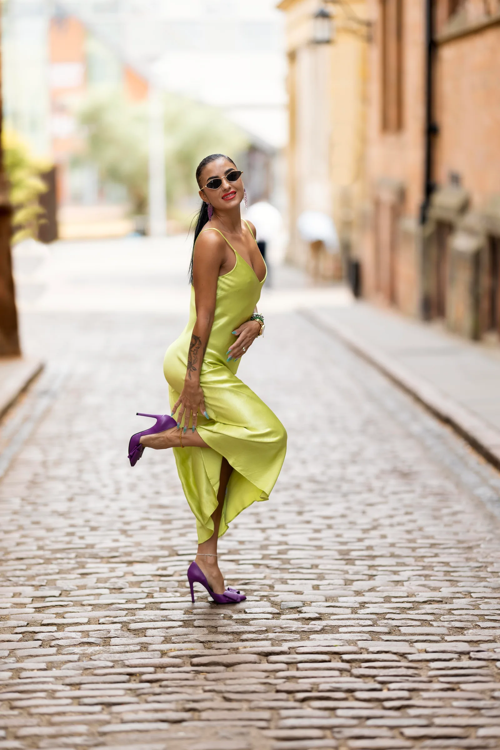 Woman in yellow satin dress mid-stride on a cobblestone street, one leg kicked up