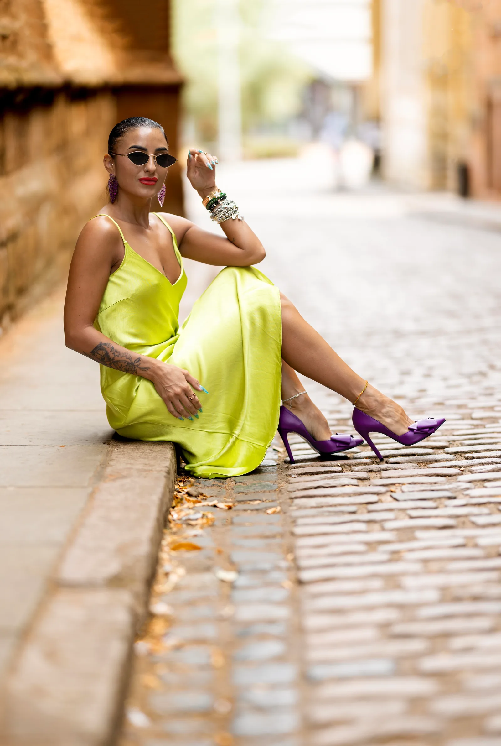 Woman in yellow satin dress sitting on a cobblestone street, legs extended, purple heels
