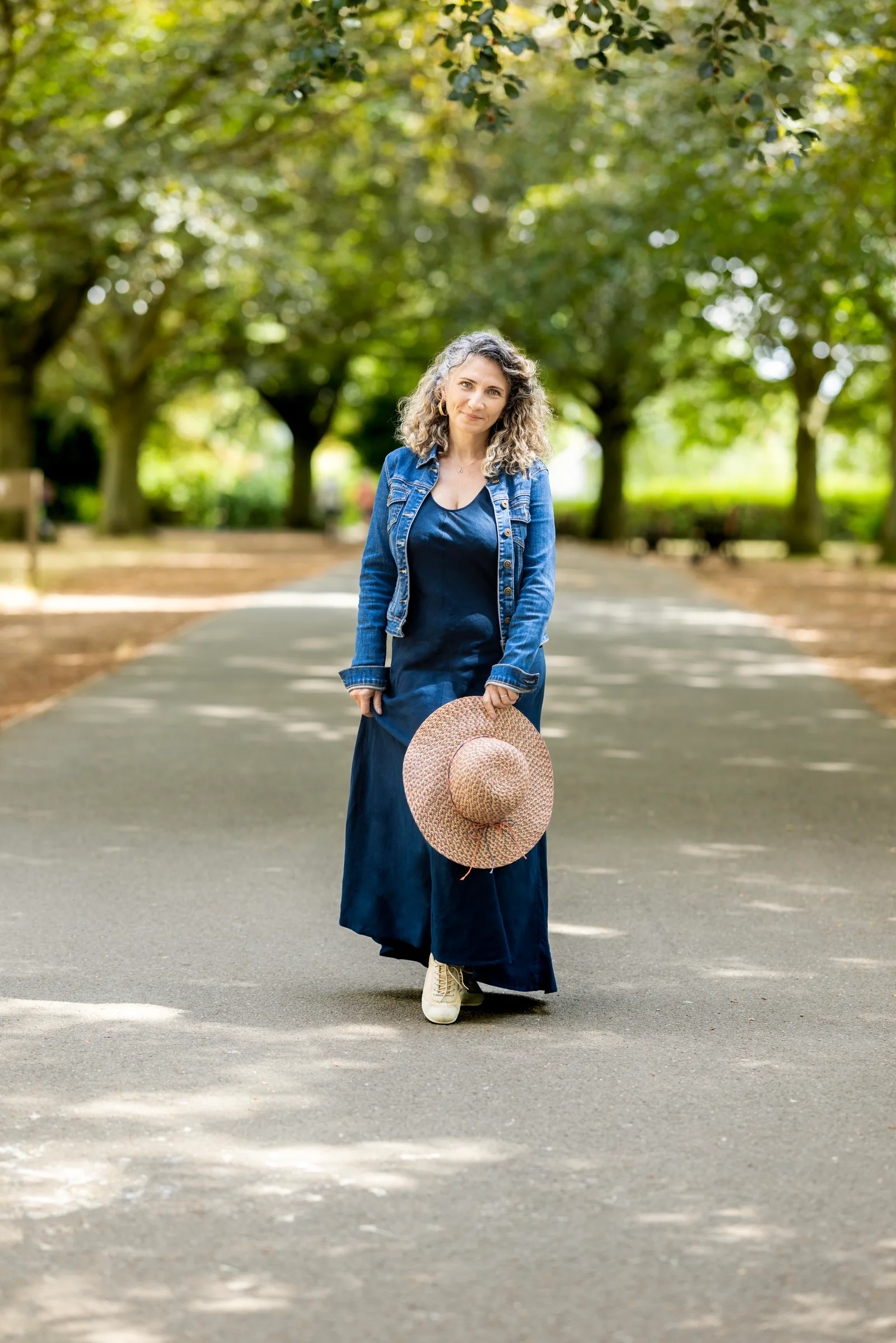 Woman in navy dress and denim jacket walking a park path, holding a straw hat