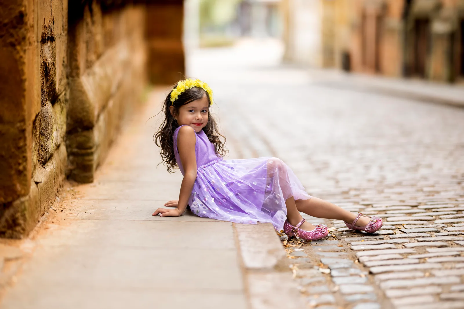 Young girl in a lilac tulle dress sitting on a cobblestone street curb