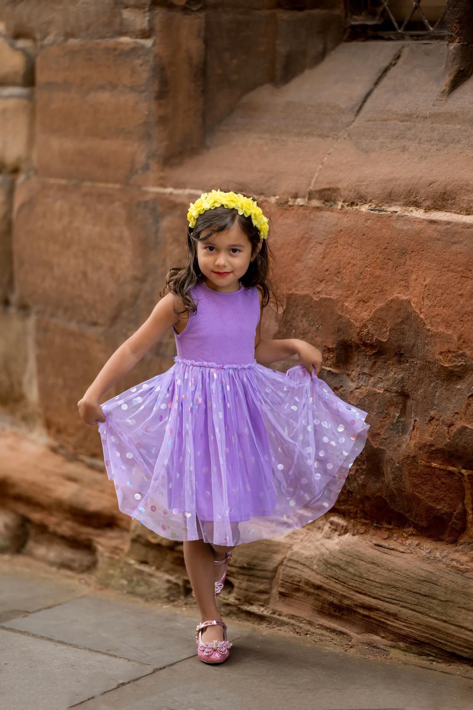 oung girl in a purple tulle dress and flower crown twirling beside a sandstone wall