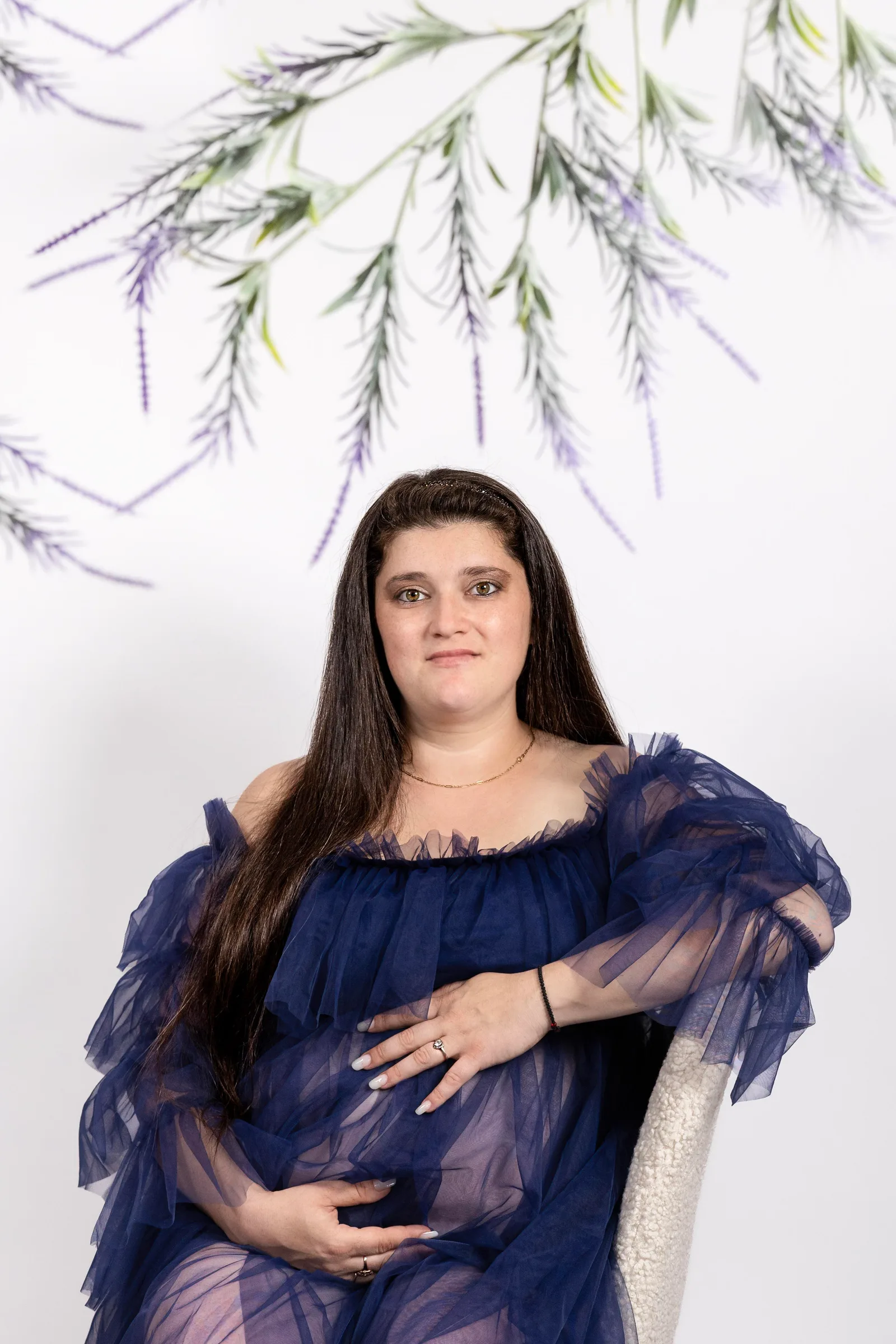 Pregnant woman in sheer navy tulle gown, seated and cradling her belly beneath lavender greenery