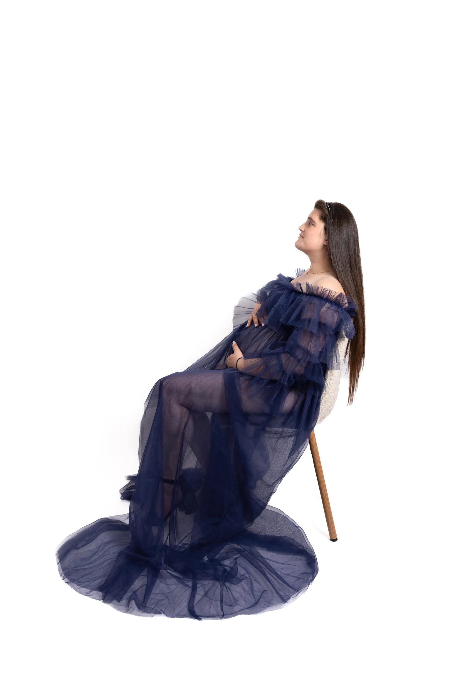 Pregnant woman in sheer navy tulle gown, seated in profile on a wooden stool